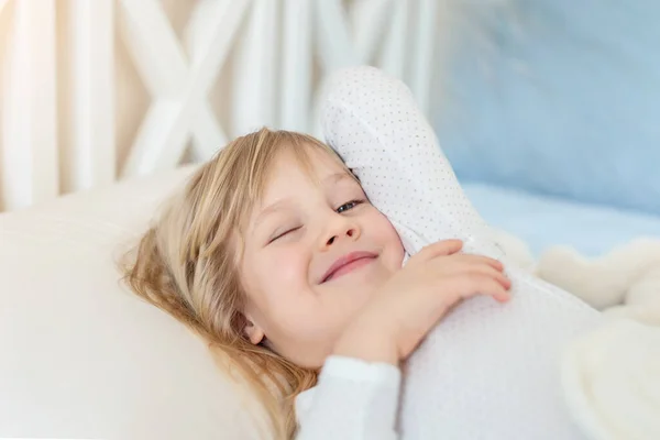 Cute adorable caucasian blond little toddler kid girl lying in bed on white pillow. Adorable sweet happy smiling child stretching wake up early morning for kindergarten or school. Childhood concept — Stock Photo, Image