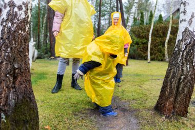 Anne ve sevimli beyaz kardeşleri açık sarı yağmurluk ve çizmelerle bahçedeki soğuk bahar yağmurlu günlerinde evlerinin arka bahçesinde oynuyorlar. Tasasız çocukluk kavramı. Kötü hava