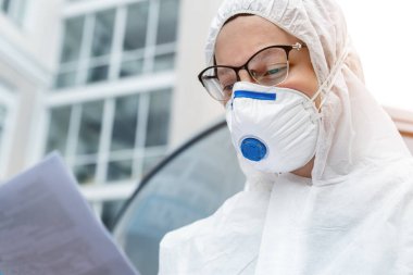 Portrait of tired exhausted female doctor, scientist or nurse wearing face mask and biological hazmat ppe suit reading who treatment protocol or research outdoor. Coronavirus covid-19 outbreak danger. clipart