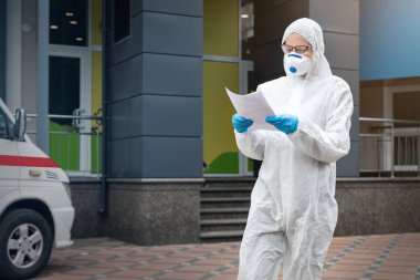 Yüz maskesi takan yorgun kadın doktor, bilim adamı ya da hemşire portresi ve dışarıda kimi tedavi ettiğini gösteren biyolojik tehlikeli madde kıyafeti. Coronavirus covid-19 salgını tehlikesi.
