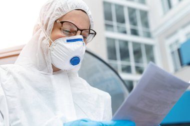 Yüz maskesi takan yorgun kadın doktor, bilim adamı ya da hemşire portresi ve dışarıda kimi tedavi ettiğini gösteren biyolojik tehlikeli madde kıyafeti. Coronavirus covid-19 salgını tehlikesi.