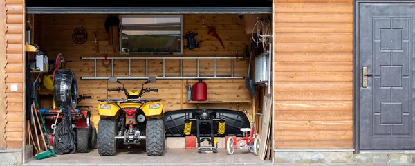 Facade front view open door ATV quad bike motorcycle parking messy garage building with wooden siding at home driveway backyard and lawn path. House warehouse for tools and equipment . Wide panoramic — Stock Photo, Image