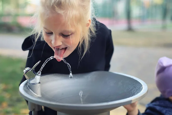 Schattig schattig blank blond klein dorstig schoolmeisje drinkwater drinken uit openbare drinkbare fontein kraan in stadspark op heldere hete zomerdag buiten. Uitdroging van kinderen tijdens het warmteseizoen — Stockfoto