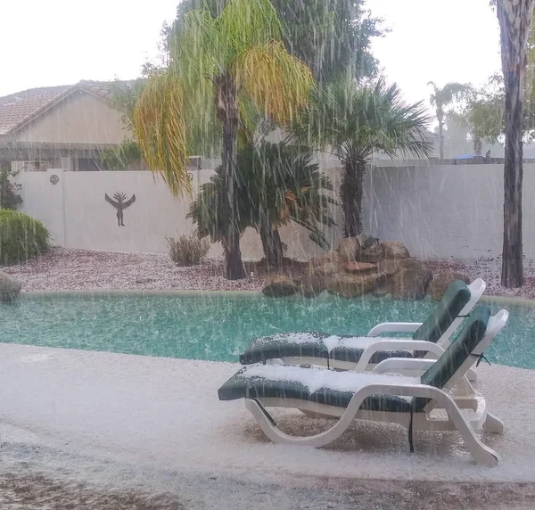 Heavy hail storm covers the lounge chairs and ground.  It also splashes into the pool. In Glendale, Maricopa County, Arizona USA