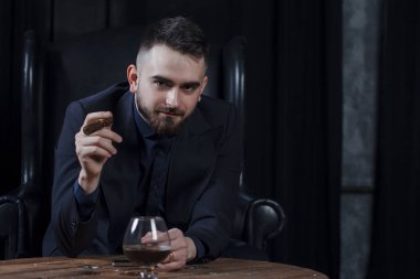 Takım elbiseli emin yakışıklı bir puro ve bir bardak viski tutarak ve kapalı deri sandalyede otururken kameraya bakıyor