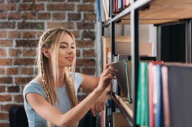 Smiling young woman choosing books on bookshelves in library  clipart