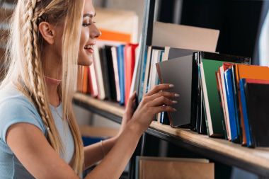 Smiling young woman choosing book on bookshelf in library  clipart
