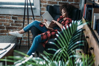 Kitap ve dijital tablet ile sandalye üzerinde oturan ve eğitim çekici genç Afro-Amerikan öğrenci