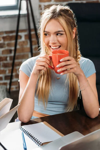 Młoda uśmiechający się kaukaski kobieta pije kawę siedząc przy stole — Zdjęcie stockowe