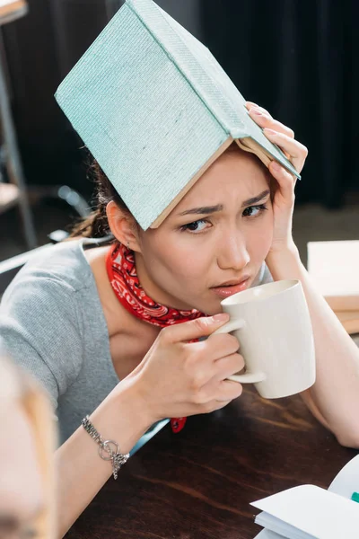 Портрет молодої втомленої азіатки, що п'є каву і тримає книгу — стокове фото