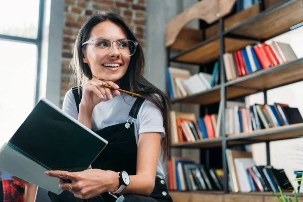 Junge lächelnde kaukasische Frau hält Notizbuch in der Hand und blickt in die Bibliothek — Stockfoto