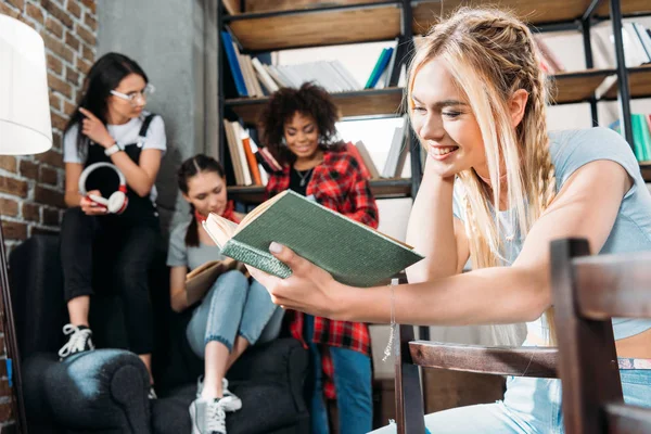 Біла дівчина читає книгу, а її друзі сидять на дивані в домашній бібліотеці — стокове фото