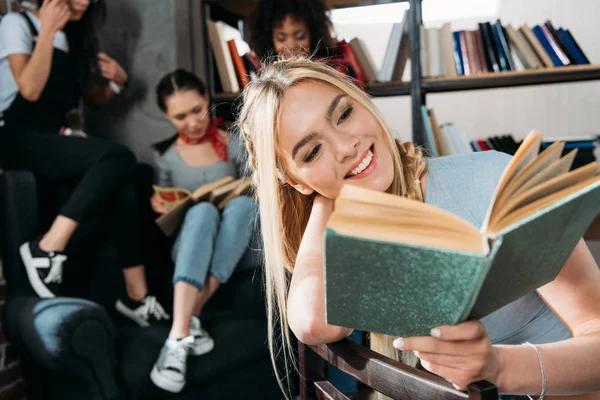 Καυκάσιος κορίτσι ανάγνωση του βιβλίου ενώ τους φίλους της να κάθεται στον καναπέ στο σπίτι βιβλιοθήκη — Φωτογραφία Αρχείου