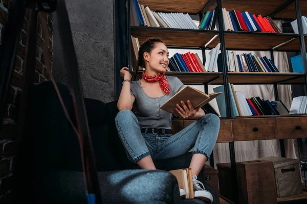 Sorrindo jovem estudante bonita sentada na cadeira com livro e olhando para longe — Fotografia de Stock