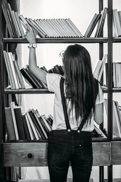 Black and white photo of young woman standing near bookshelves and choosing books