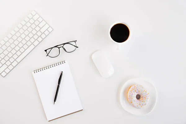 Top view of blank notebook with pen, eyeglasses, keyboard, computer mouse, cup of coffee and tasty doughnut at workplace — Stock Photo, Image