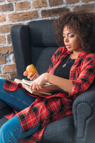Сосредоточена молодая женщина, сидящая в кресле и занимающаяся с книгами во время еды яблока — стоковое фото
