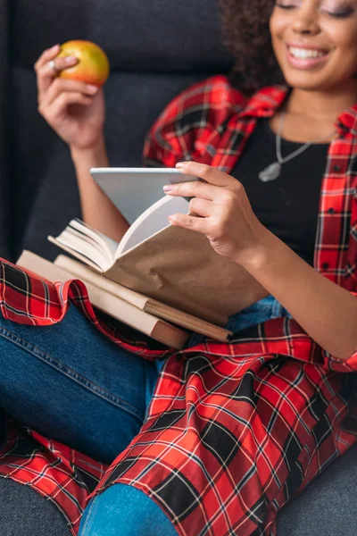 Улыбающаяся молодая женщина сидит в кресле с книгами и с помощью цифрового планшета — стоковое фото