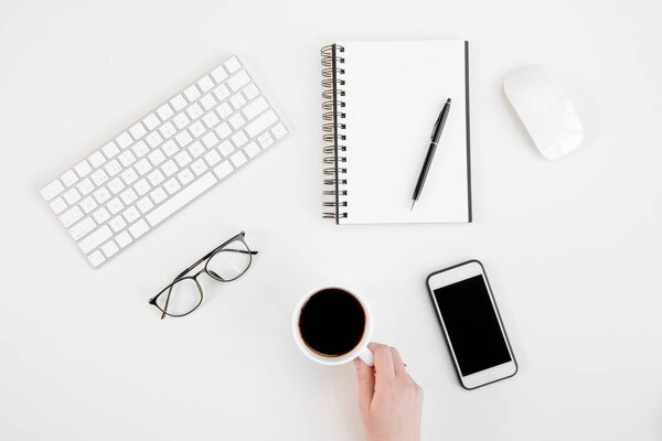 Top view of person holding cup of coffee, smartphone with blank screen, eyeglasses, keyboard and notebook with pen at workplace