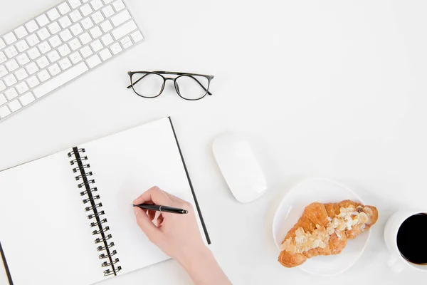Top view of person writing in notebook with pen, eyeglasses, keyboard and coffee cup with croissant at workplace — Stock Photo, Image