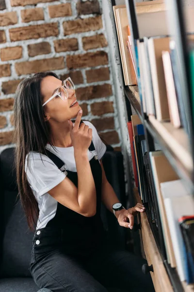 Attraktive junge Studentin mit Brille, die Bücher auswählt und Bücherregale betrachtet Stockfoto