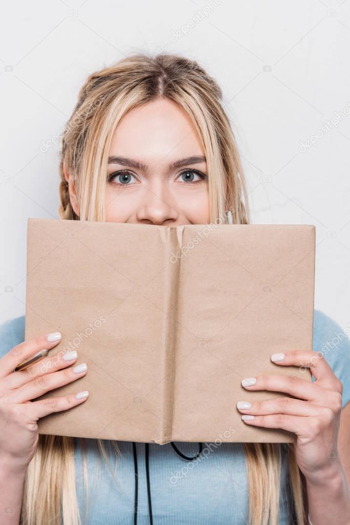 young blonde woman holding book in front of face and looking at camera