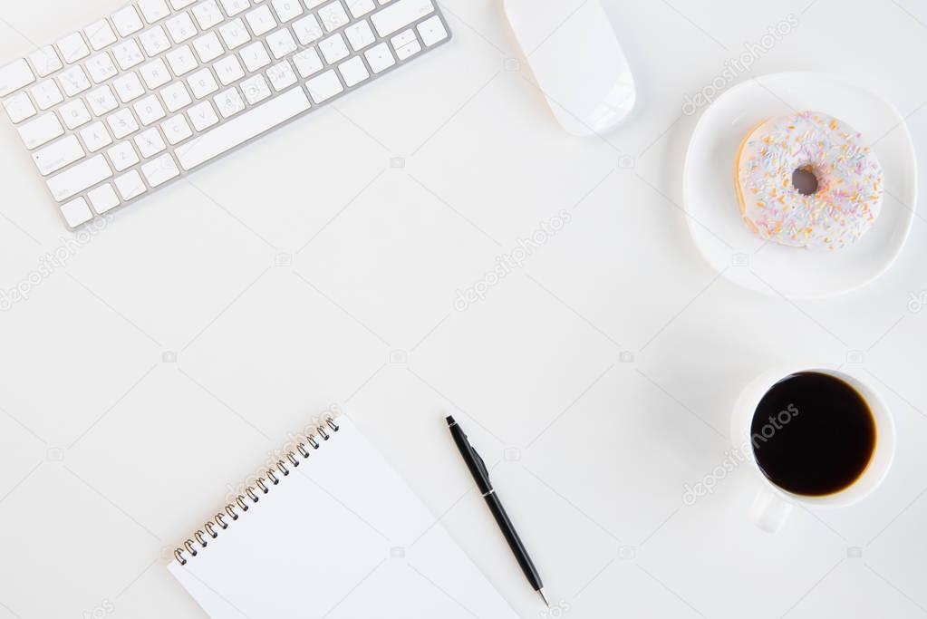 Top view of doughnut with cup of coffee, blank notebook with pen, keyboard and computer mouse at workplace