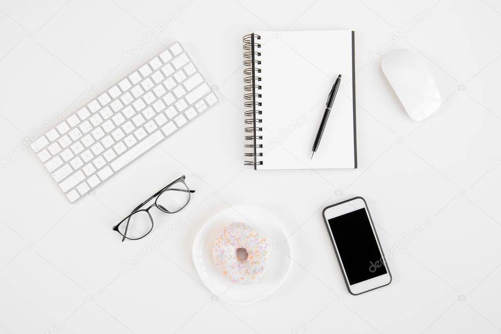 Top view of smartphone with blank screen, blank notebook with pen, eyeglasses, keyboard, computer mouse and tasty doughnut at workplace