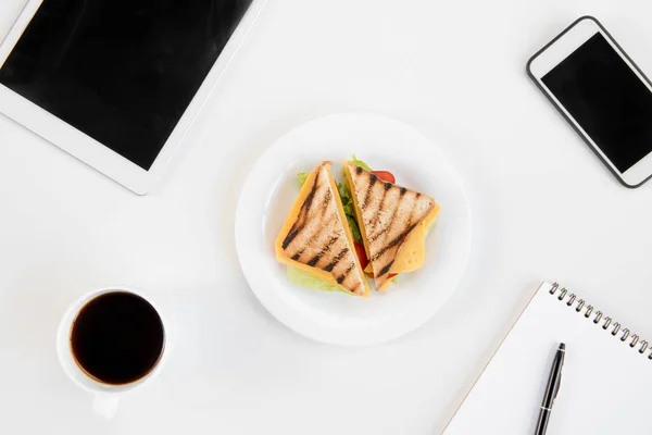 Vista superior de sabrosos sándwiches en el plato, taza de café, portátil con pluma, smartphone y tableta digital en el lugar de trabajo —  Fotos de Stock