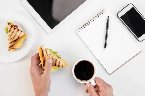 Visão superior da pessoa comendo sanduíches e bebendo café no local de trabalho com eletrônicos — Fotografia de Stock