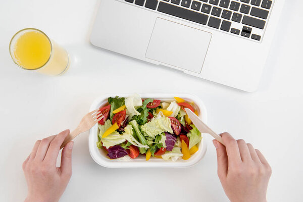 Обрезанный снимок человека, который ест свежий салат и пьет апельсиновый сок на рабочем месте с ноутбуком

