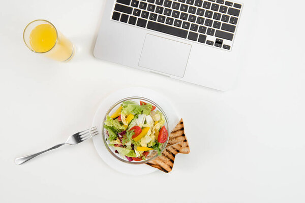 Вид сверху ноутбука, апельсиновый сок в стакане и свежий здоровый салат с тостами на рабочем месте
