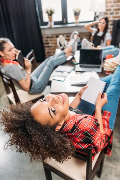 Attractive young african american student sitting at table with classmates and smiling at camera — Stock Photo, Image