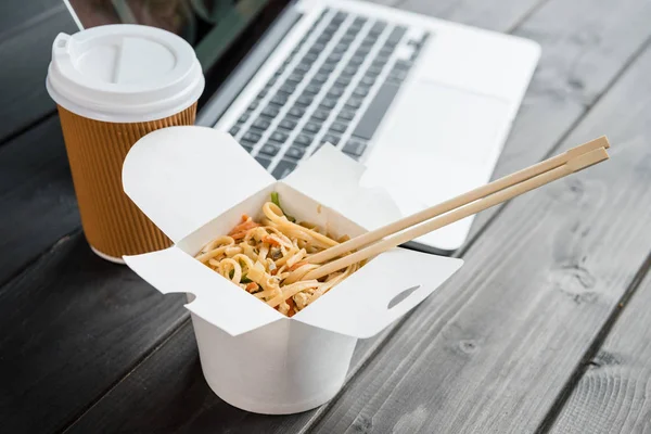 Close up de macarrão com pauzinhos e café para ir com laptop em mesa de madeira — Fotografia de Stock