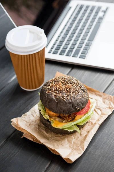 Close up of burger with coffee to go and laptop on wooden tabletop — Stock Photo, Image