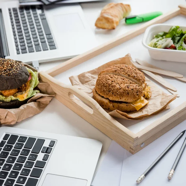 Close up of tasty burgers with fresh salad and bread loaf with laptops on tabletop — Stock Photo, Image