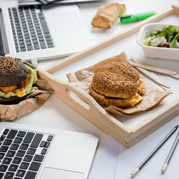 close up of tasty burgers with fresh salad and bread loaf with laptops on tabletop