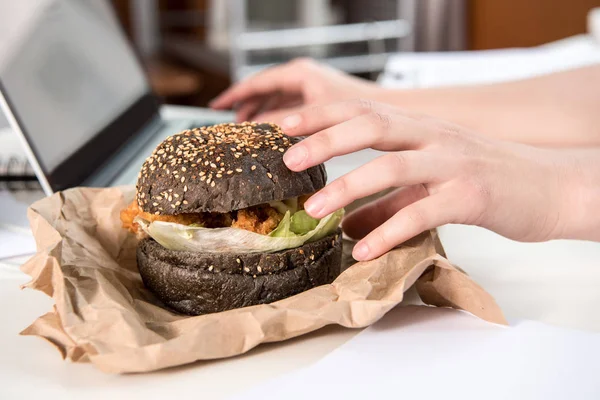 Close up of tasty burger with black bun on tabletop at workplace — Stock Photo, Image