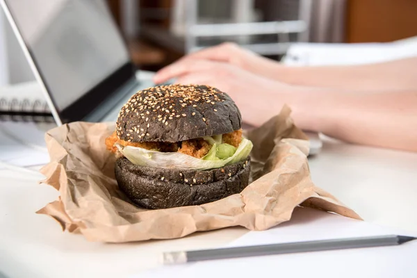 Close up of tasty burger with black bun on tabletop at workplace — Stock Photo, Image
