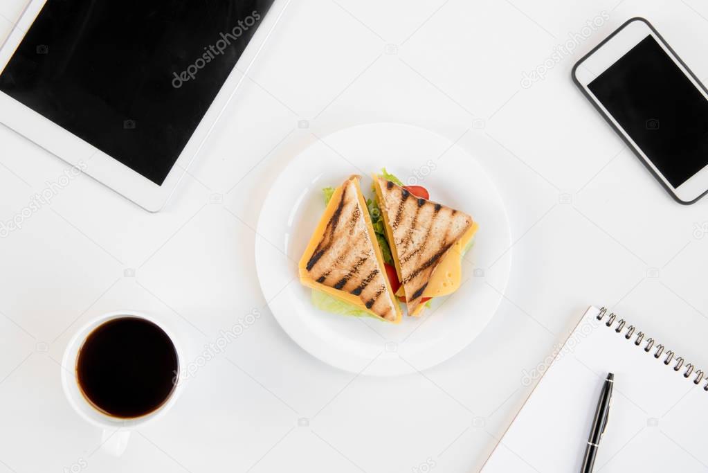Top view of tasty sandwiches on plate, cup of coffee, notebook with pen, smartphone and digital tablet at workplace