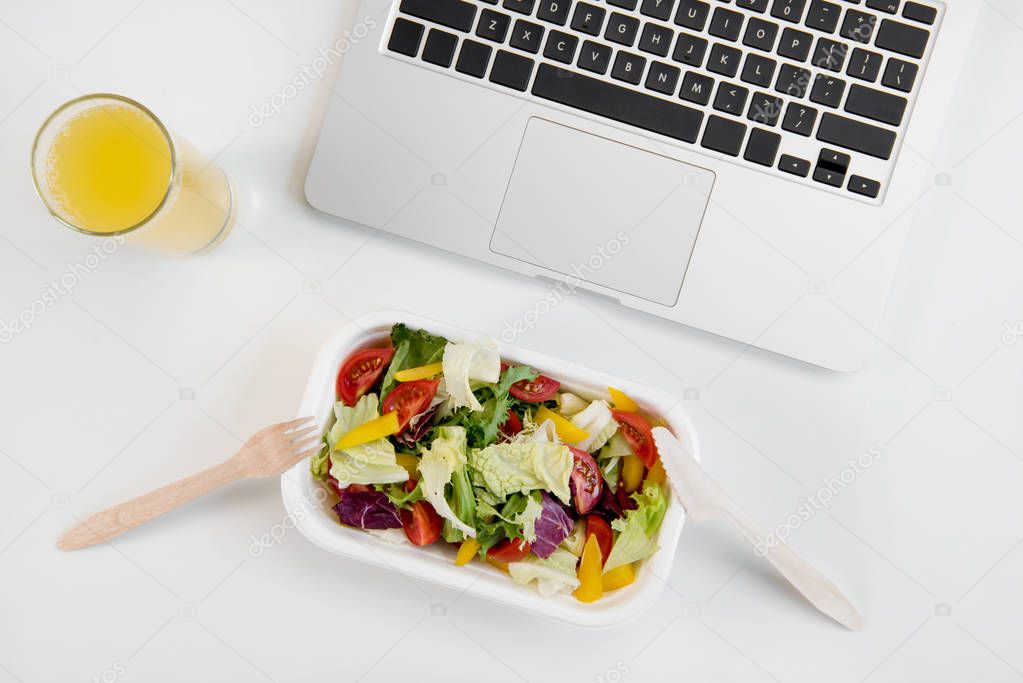Top view of laptop, orange juice in glass and fresh salad in lunch box with plastic utensils at workplace