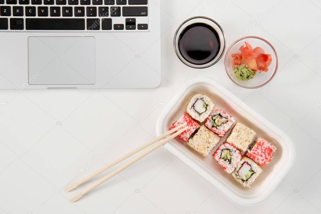 Top view of open laptop and sushi rolls with wasabi and soy sauce at workplace