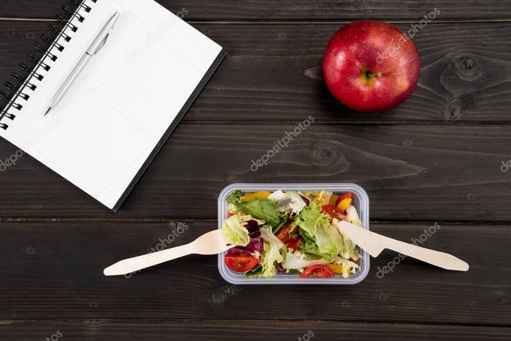 Top view of natural salad with red apple and notebook on wooden surface