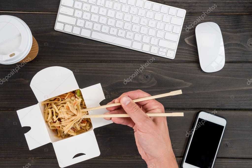 hand holding chopsticks with noodle and keyboard with smartphone on wooden tabletop
