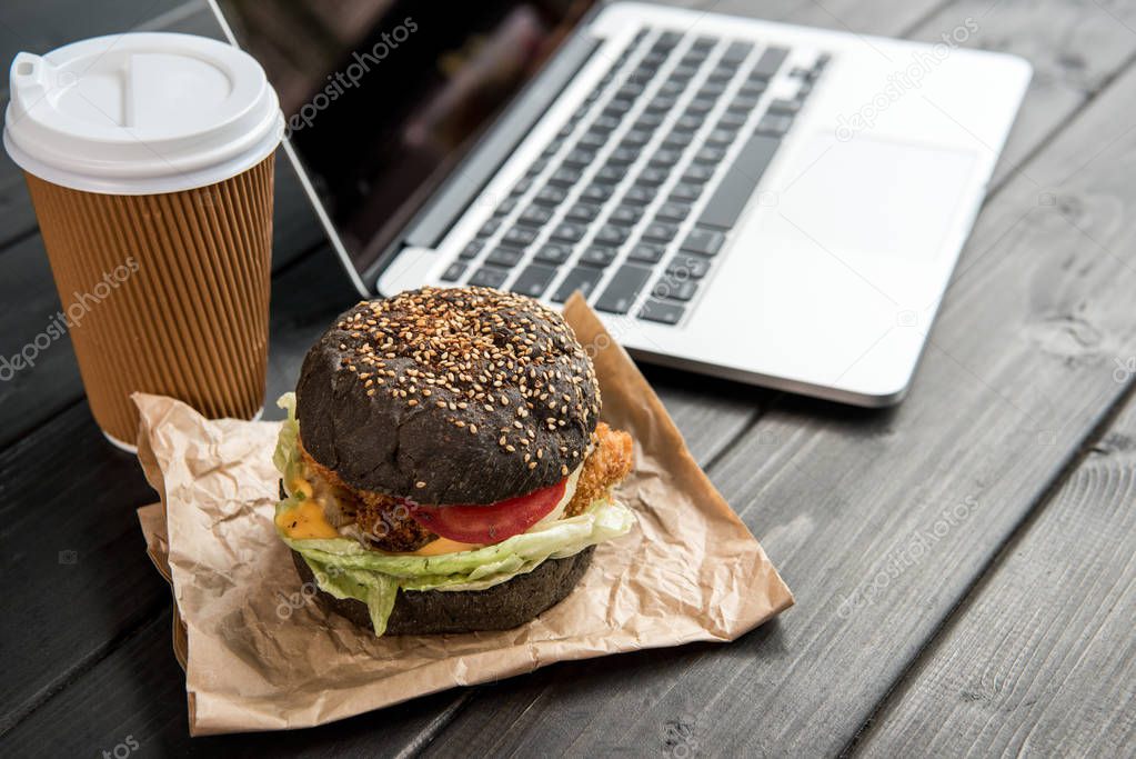 close up of burger with coffee to go and laptop on wooden tabletop