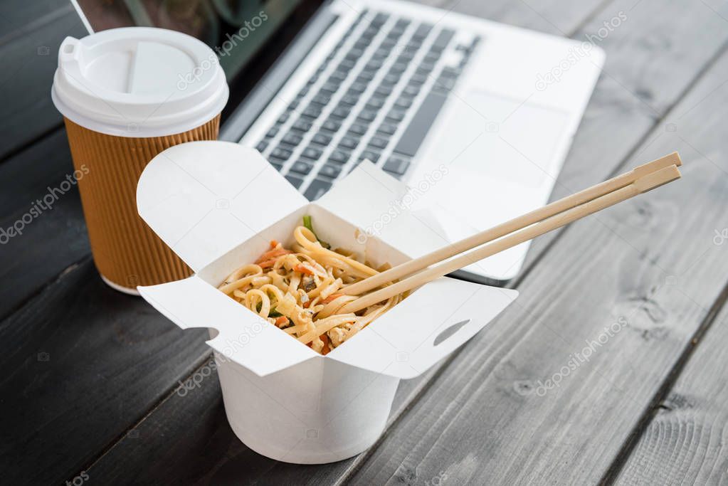 close up of noodle with chopsticks and coffee to go with laptop on wooden tabletop