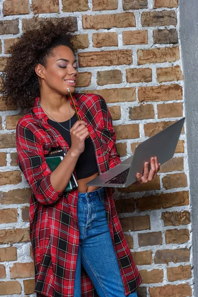 Young african american woman smiling while using laptop and holding notebooks — Stock Photo