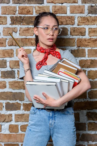 Retrato de pensativo asiático estudiante sosteniendo libros y lápiz - foto de stock