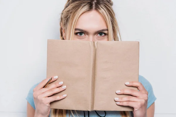 Jovem loira segurando livro na frente do rosto e olhando para a câmera — Fotografia de Stock