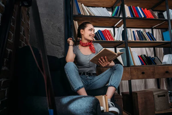 Sonriente joven hermosa estudiante sentado en la silla con libro y mirando hacia otro lado - foto de stock